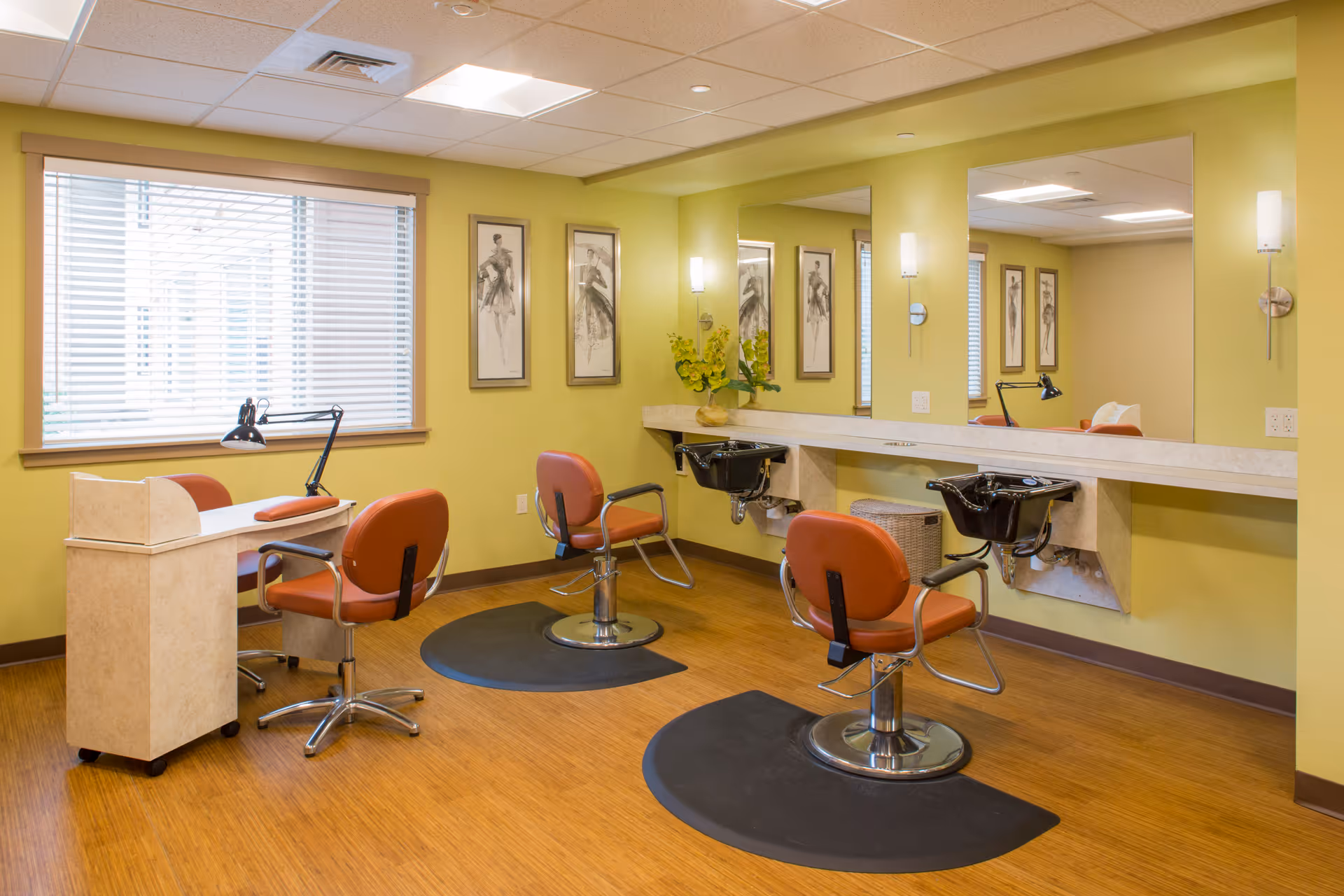Interior view of a salon area in a senior living facility with two orange salon chairs in front of black wash basins mounted on a beige counter. There is a manicure table with an orange chair and a desk lamp near a window with blinds. The walls are painted light green and decorated with framed artwork. The floor is wooden with black mats under the salon chairs.