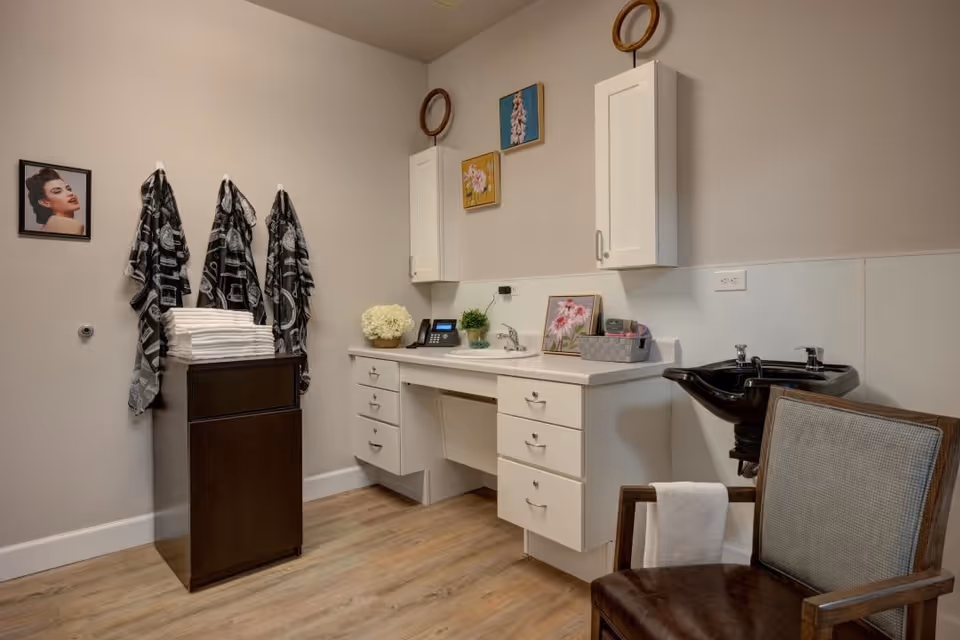 A clean and organized hair washing station in a senior living facility with a black shampoo sink, a cushioned chair with a towel draped over the armrest, a countertop with drawers, a small sink, decorative flowers, and three black and white patterned towels hanging on hooks next to a stack of white towels on a dark wooden cabinet. The walls are light-colored with framed pictures and two white wall cabinets.