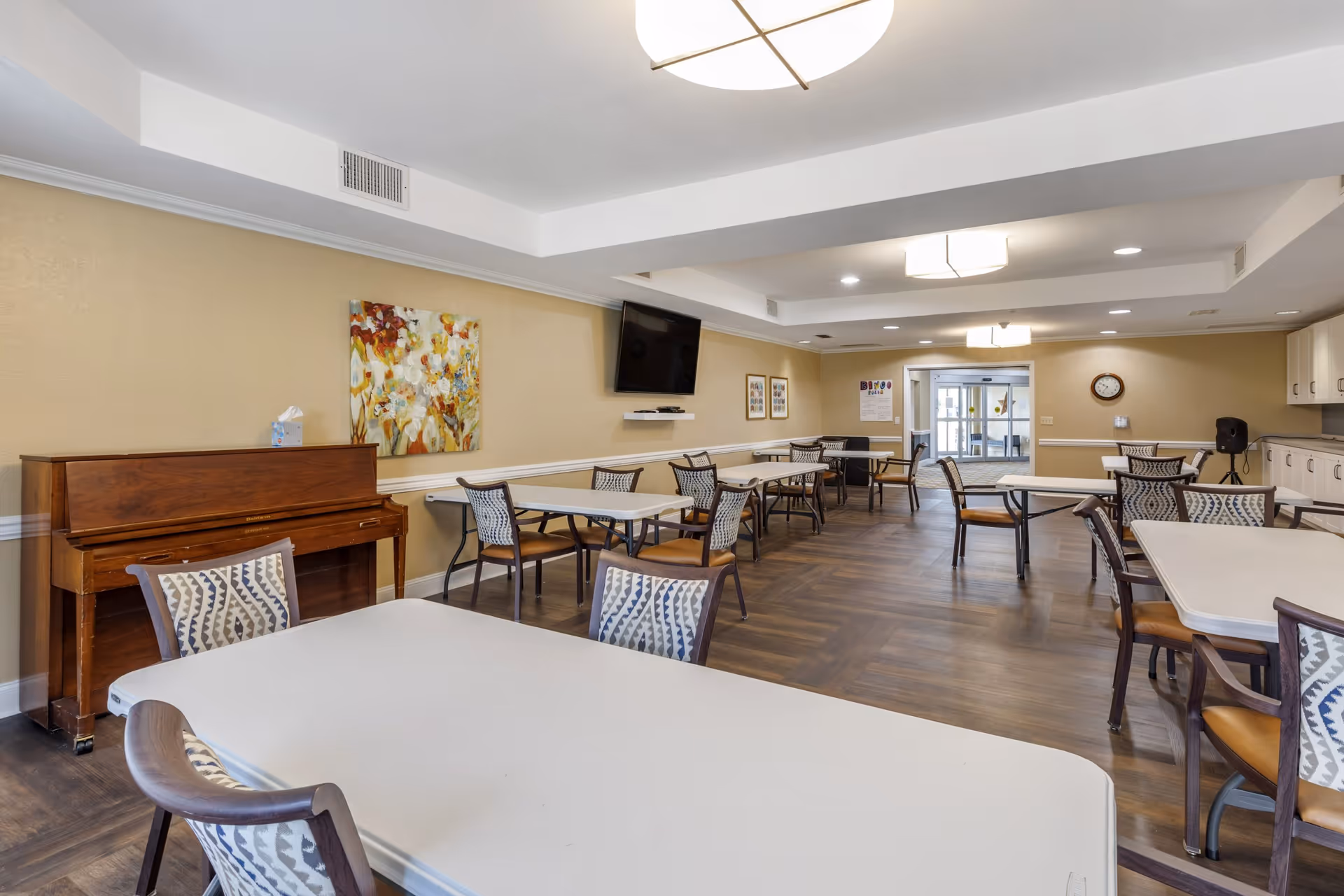 Spacious communal dining room with multiple tables and chairs, a piano, wall-mounted TV, and decorative artwork.