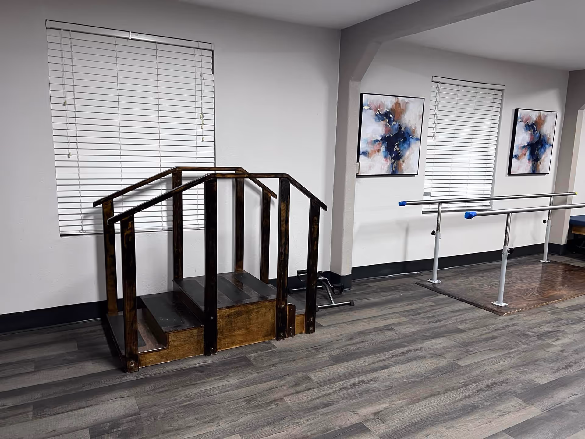 An interior physical therapy room with wooden practice stairs and parallel bars in front of windows with blinds and abstract artwork.