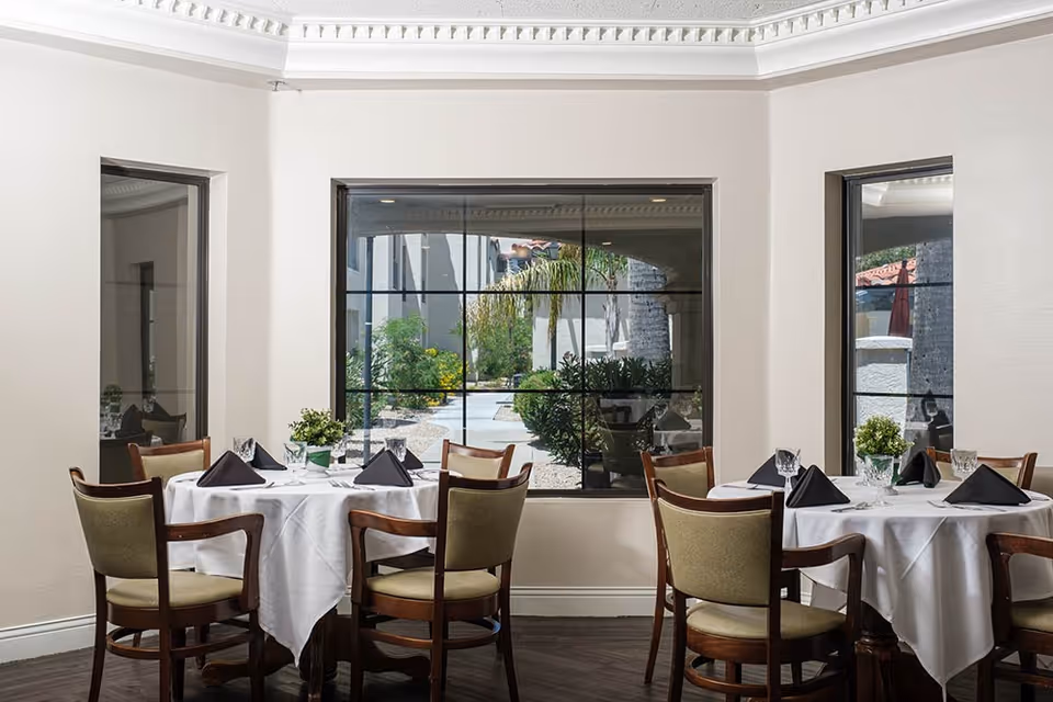 Dining area with two round tables covered with white tablecloths, each set with four chairs. Tables are set with black folded napkins, glassware, and small green plants as centerpieces. Large windows behind the tables show an outdoor garden area with plants and a pathway.