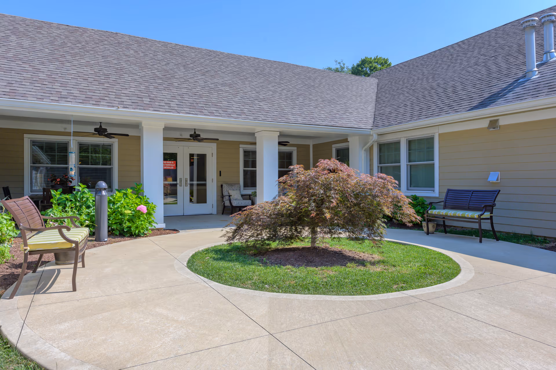 Outdoor courtyard area of a senior living facility with a circular concrete walkway surrounding a small tree in the center. The building has beige siding, white trim, and a shingled roof. There are benches with cushions and some green plants near the building entrance with double glass doors.