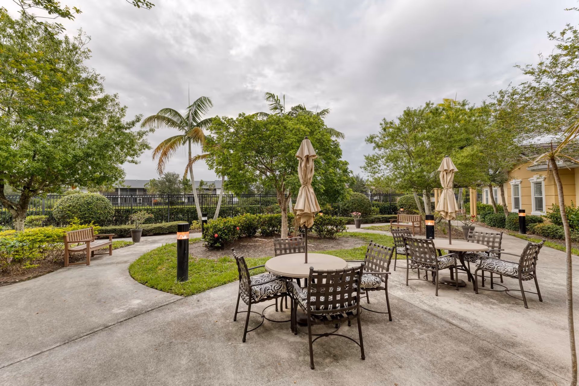 Outdoor patio area with round tables and chairs, each table having a closed umbrella in the center. The patio is surrounded by green trees, bushes, and a few benches along the concrete pathways. The sky is cloudy.
