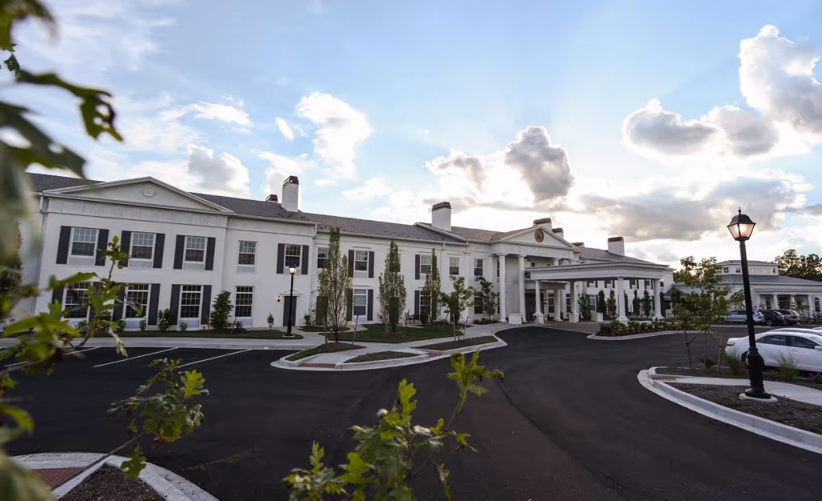 Front exterior of a large white two-story senior living building with columns, a circular driveway, lampposts, and landscaped grounds under a cloudy sky.