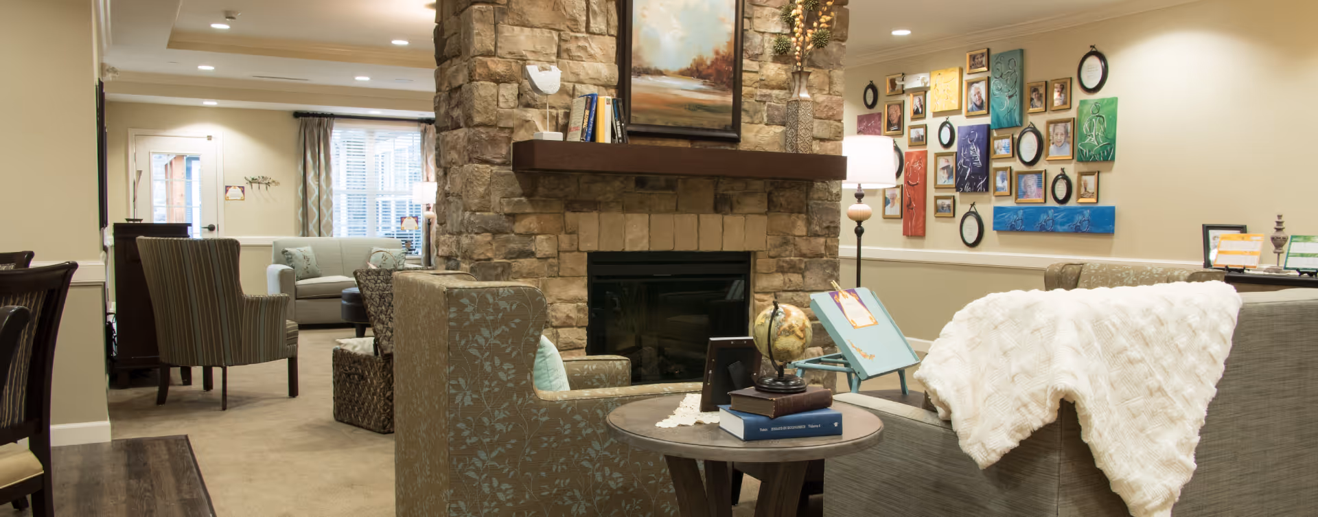 A cozy living room area in a senior living facility featuring a stone fireplace with a painting above it. The room has comfortable armchairs and sofas, a side table with books and a globe, and a wall decorated with various framed pictures and artwork. Soft lighting and neutral tones create a warm and inviting atmosphere.