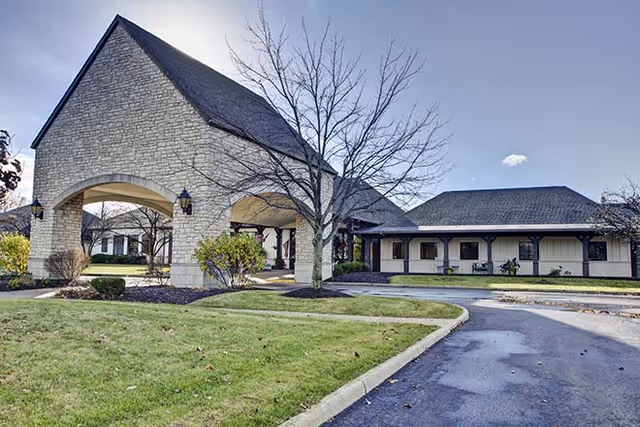 Exterior view of a senior living facility building with a stone archway entrance, a paved driveway, and landscaped grass and shrubs under a clear sky.