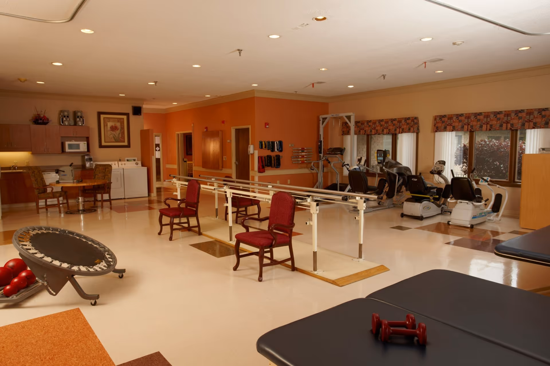 A spacious rehabilitation room with exercise equipment including a small trampoline, red dumbbells on a padded table, parallel bars with red chairs, and various cardio machines near windows. The room has beige and orange walls, a kitchenette area with a microwave and washing machines, and a table with chairs.