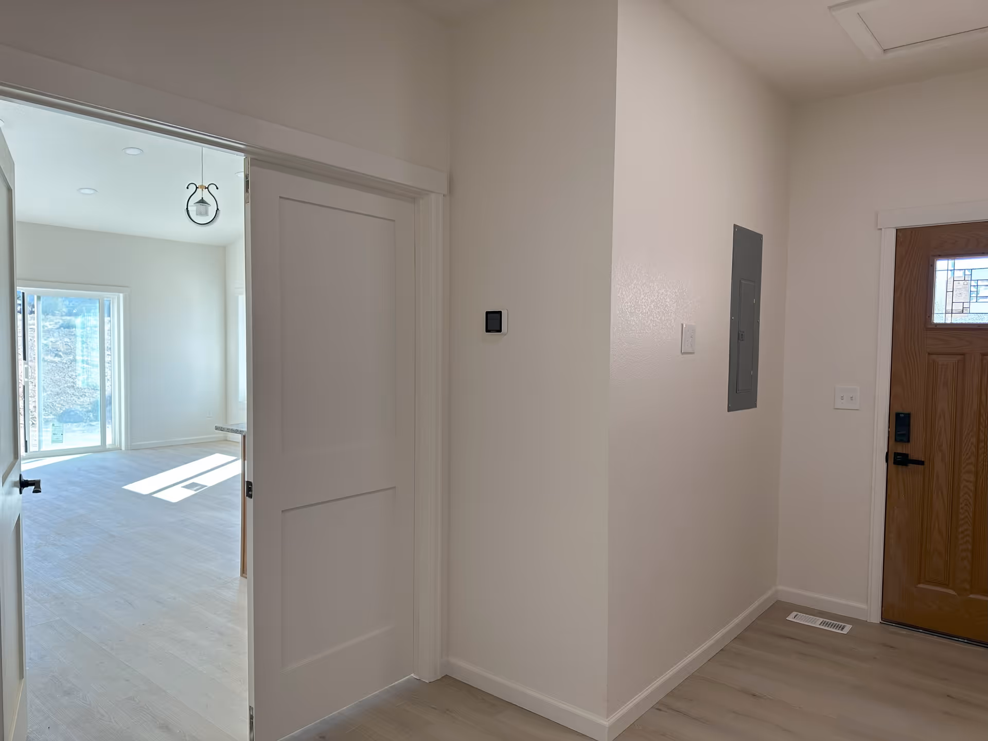 Interior view of a hallway in a senior living facility with light-colored walls and flooring. A wooden front door with a decorative glass panel is visible on the right. To the left, an open doorway leads to a bright room with large windows letting in natural light.