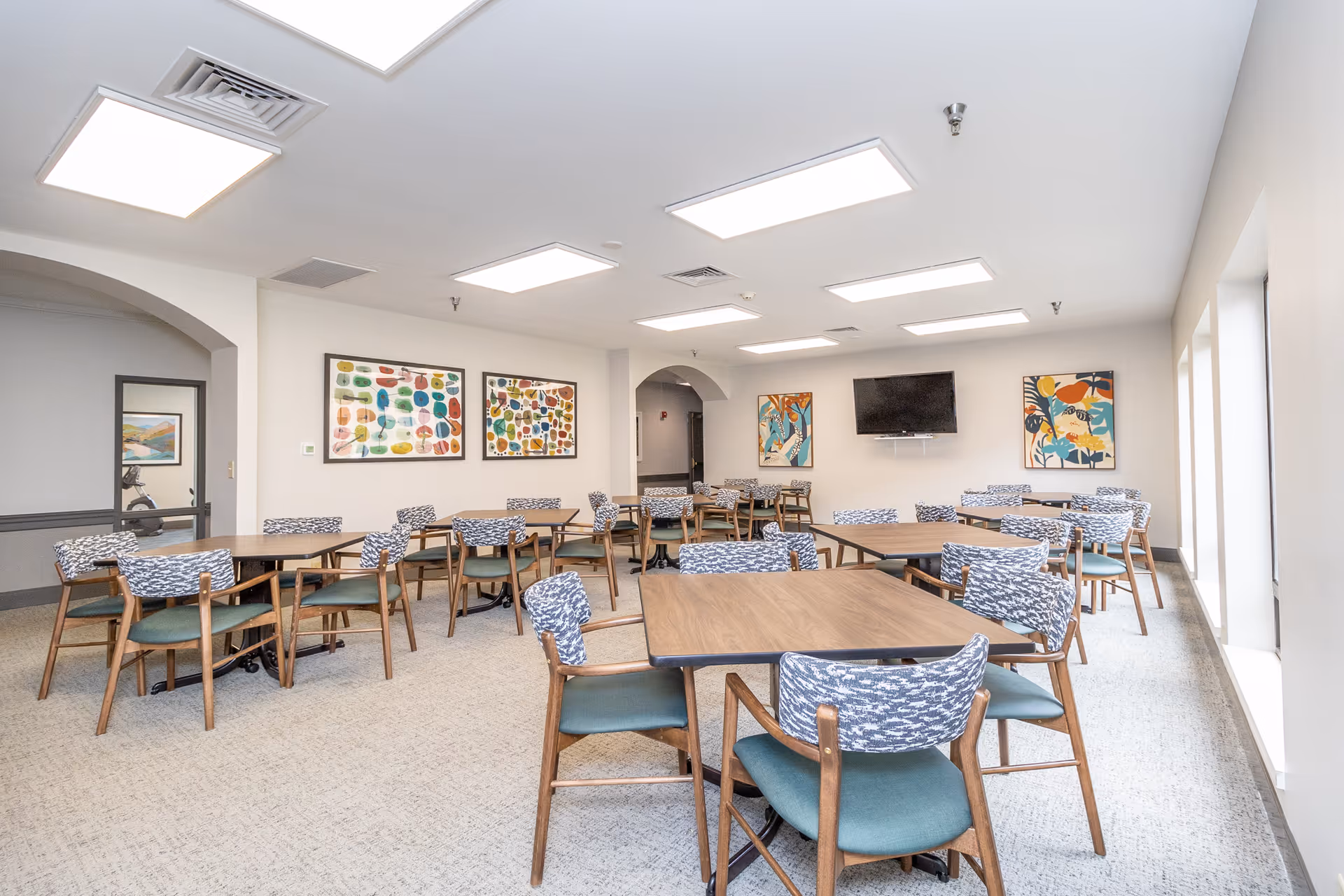 A bright and spacious dining room with multiple wooden tables and chairs featuring patterned upholstery. The room has large windows on one side allowing natural light to fill the space. Colorful abstract artwork is displayed on the walls, and a flat-screen TV is mounted on one wall. The ceiling has several rectangular recessed lights.