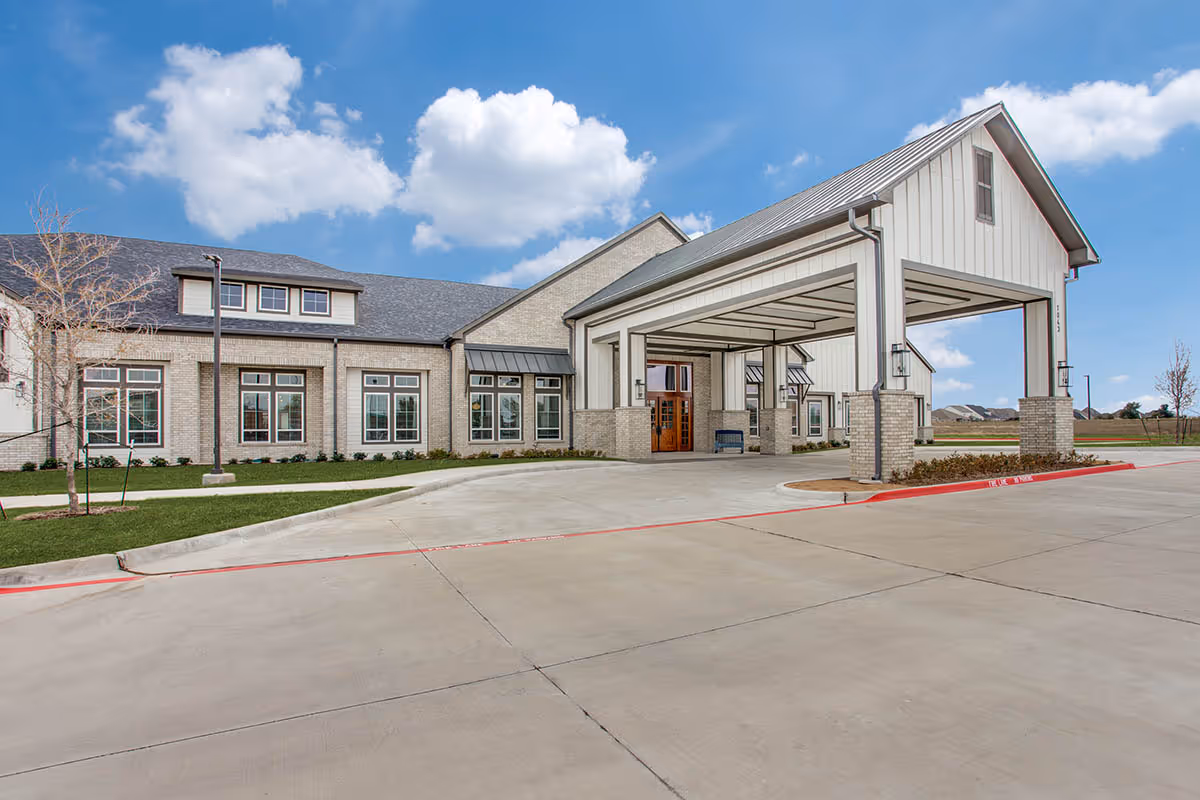 Exterior view of The Harvest Of Aledo facility showing a large covered entrance with brick and siding walls, multiple windows, a concrete driveway, and a partly cloudy blue sky.