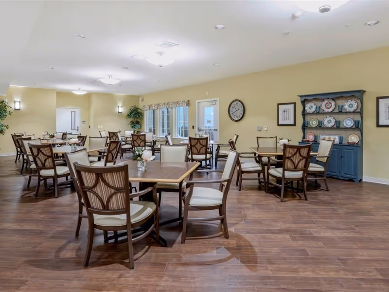 Spacious dining room with multiple wooden tables and upholstered chairs and a decorative plate hutch against a pale yellow wall.