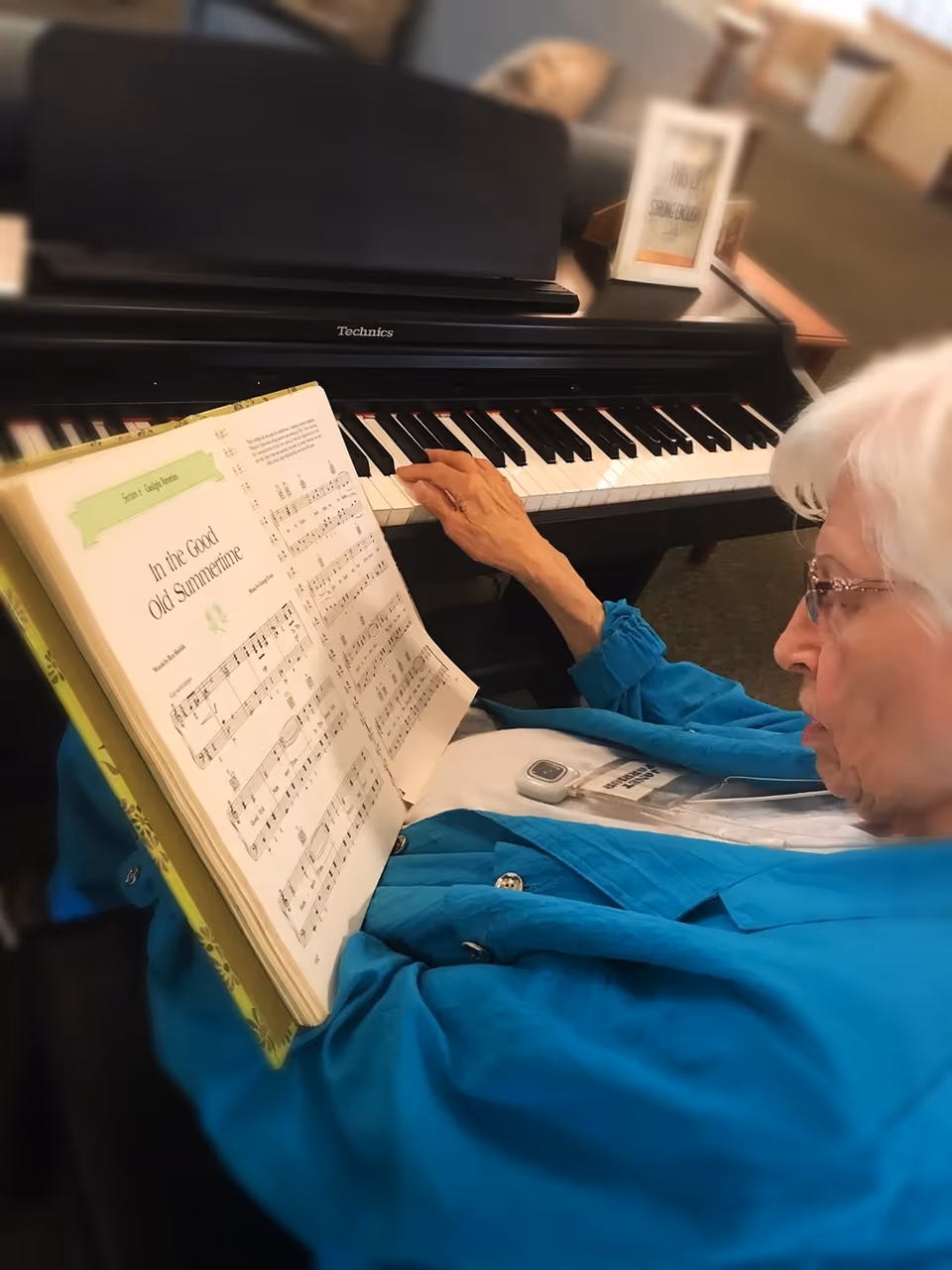 An elderly woman wearing glasses and a blue jacket is seated at a black Technics piano, reading sheet music titled 'In the Good Old Summertime' while playing the piano keys with her right hand.