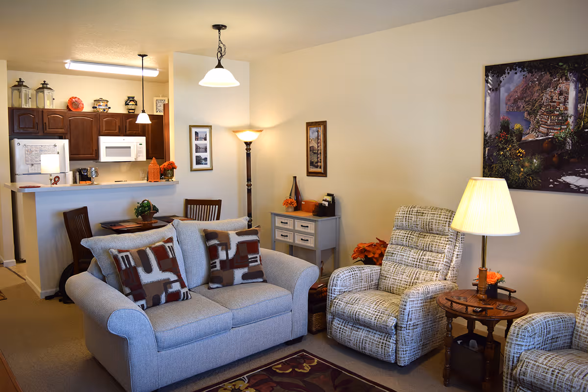 A cozy living room area with a light gray loveseat adorned with patterned pillows, a matching armchair, and a wooden side table with a lamp and small flower arrangement. Behind the living room is a small dining area with a round table and chairs, and a kitchen with wooden cabinets, a white refrigerator, and a microwave. The walls are decorated with framed pictures and a large painting. The room is warmly lit with ceiling lights and floor lamps.