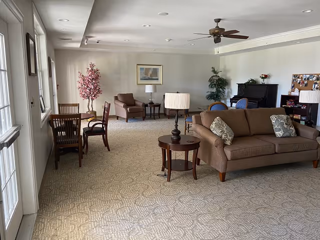 A spacious living room area with beige carpet and neutral walls. The room features a brown sofa with patterned cushions, a matching armchair, a round wooden side table with a lamp, and a small dining table with chairs near the windows. There is a ceiling fan above, a piano against the far wall, a pink artificial tree, and some framed artwork and plants decorating the space.