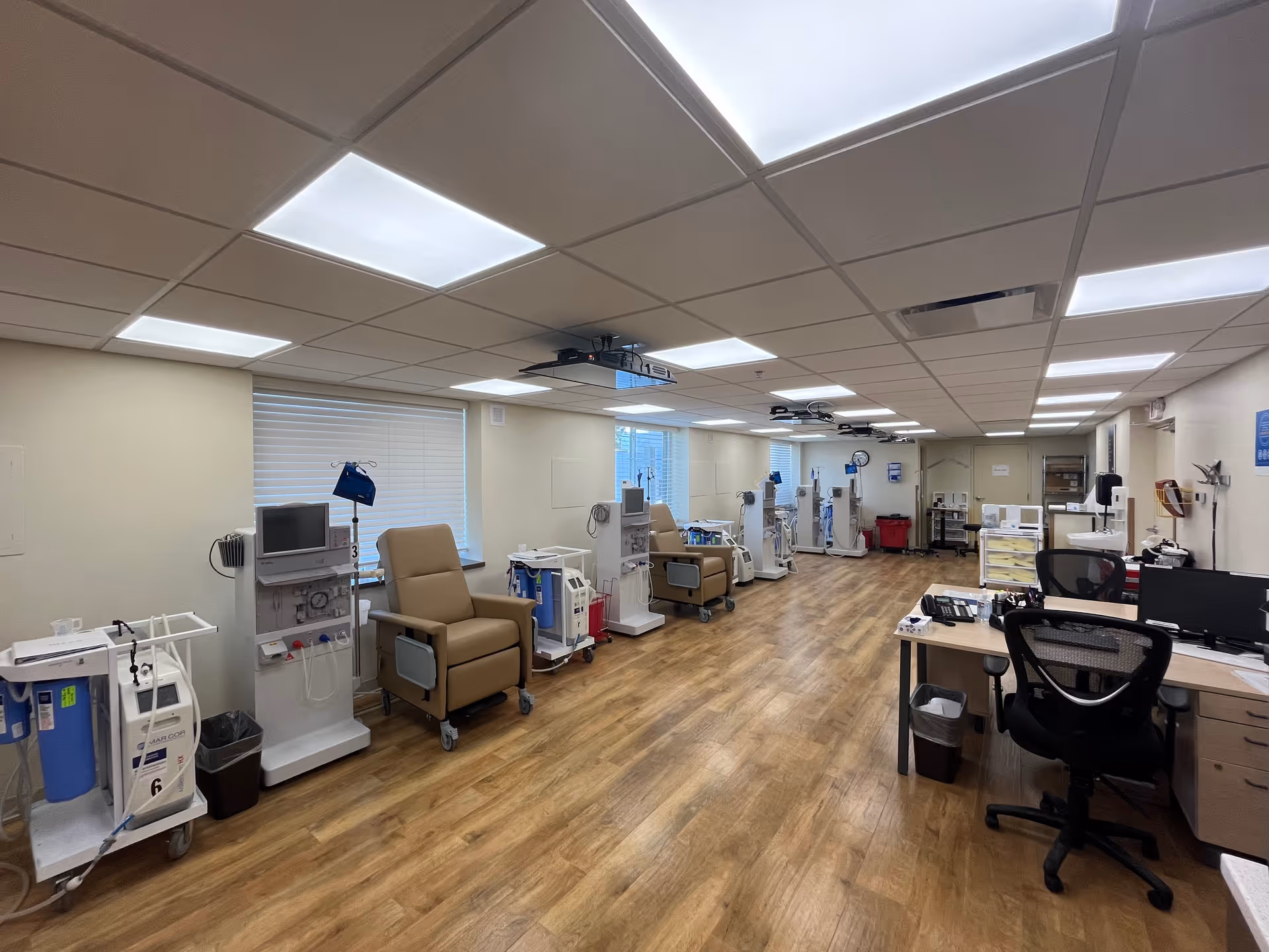 A medical treatment room with multiple dialysis machines lined up along the walls, each paired with a beige recliner chair. The room has wood-patterned flooring, white walls, and a drop ceiling with fluorescent lighting. There is a desk with office chairs and computer monitors on the right side of the room.