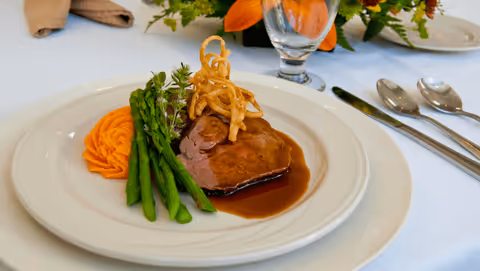 A plated meal featuring a slice of roast beef with brown gravy, garnished with crispy fried onions, served with steamed asparagus and a swirl of mashed sweet potatoes on a white plate. The plate is set on a white tablecloth with a glass of water and silverware nearby, and a floral centerpiece in the background.