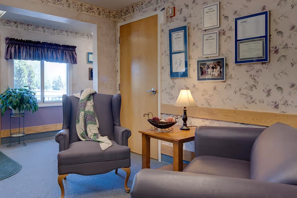 A cozy sitting area in a senior living facility with a gray armchair draped with a crocheted blanket, a gray sofa, a wooden side table with a small lamp and decorative bowl, floral wallpaper with framed certificates and photos, a wooden door, and a window with a purple valance and a potted plant on a stand.