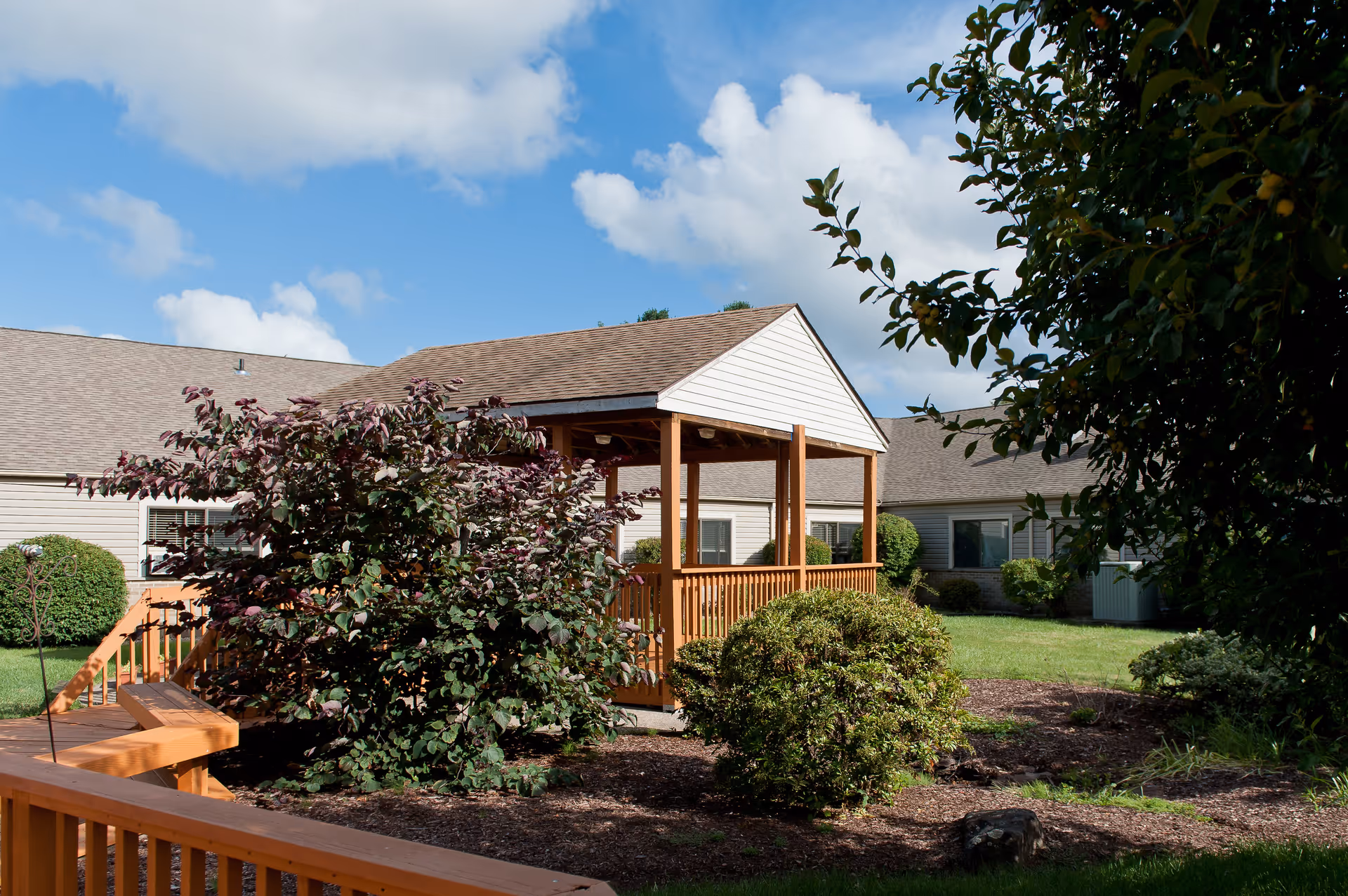 Wooden gazebo and landscaped courtyard in front of a single-story senior living building under a blue sky.
