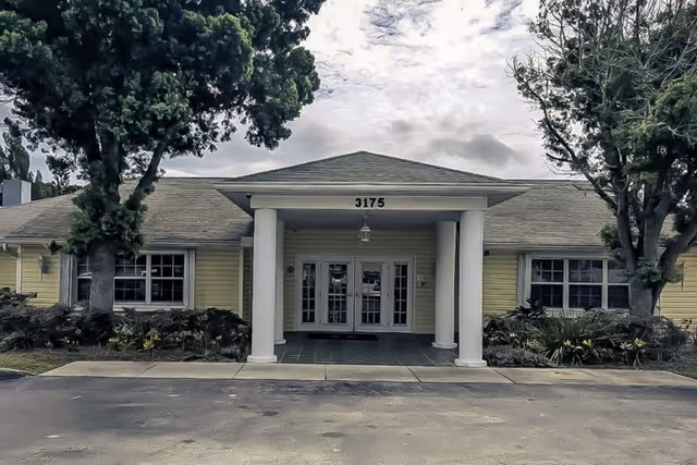 Front exterior view of a single-story building with yellow siding, white columns supporting a small portico, and the number 3175 above the entrance. There are two large trees on either side of the entrance and some landscaping with bushes and plants.