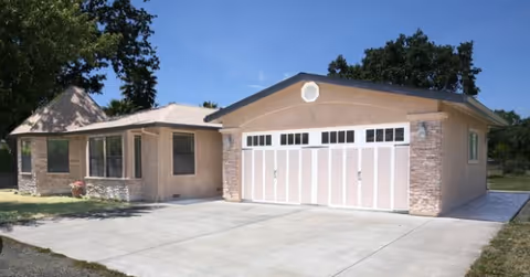 Single-story residential building with a beige exterior, stone accents, and a large white garage door under a clear blue sky. The driveway is concrete and there are trees in the background.