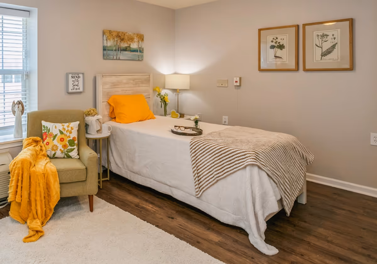 A cozy single bedroom with a bed, armchair, side table, lamp, and framed artwork on neutral walls.