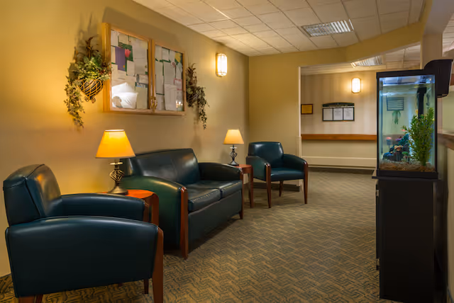 A small lounge/waiting area with green leather chairs and a sofa, side tables with lamps, a bulletin board on the wall, and an aquarium.