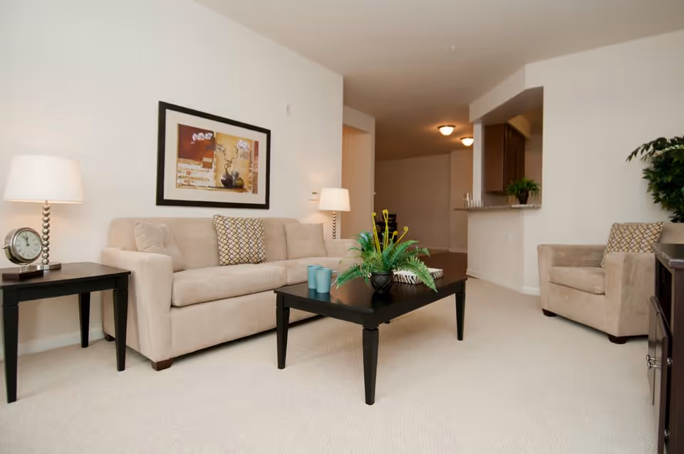 Bright living room with a beige sofa and armchair, dark coffee and side tables, framed artwork, and a potted plant on the table.