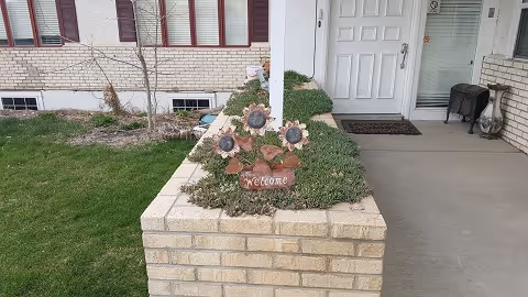 Entrance area of a residential assisted living facility with a brick planter containing a decorative welcome sign featuring sunflowers. The entrance has a white door, a doormat, and a small bench to the right. There is a grassy area and some plants around the planter.