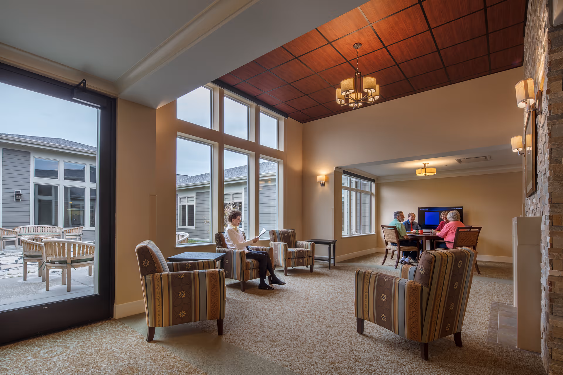 A cozy senior living facility common area with large windows letting in natural light. There are several striped armchairs arranged around small tables, a stone fireplace on the right, and a group of four seniors sitting at a table in the background watching a television. Outside the windows, there is a patio with wooden chairs and tables.