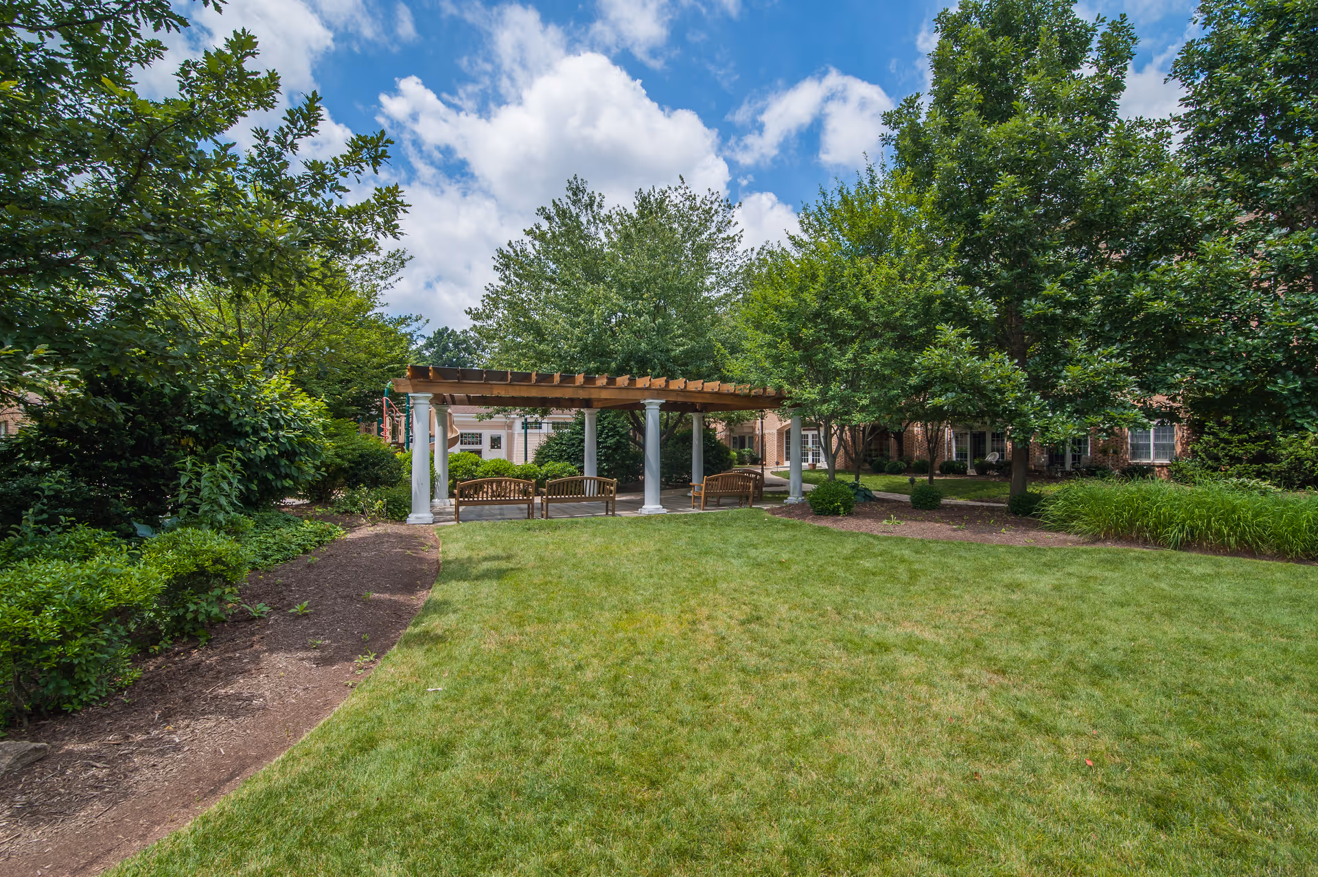 Outdoor garden area with a wooden pergola supported by white columns, surrounded by green trees and bushes, with benches underneath the pergola and a well-maintained grassy lawn in the foreground under a partly cloudy blue sky.