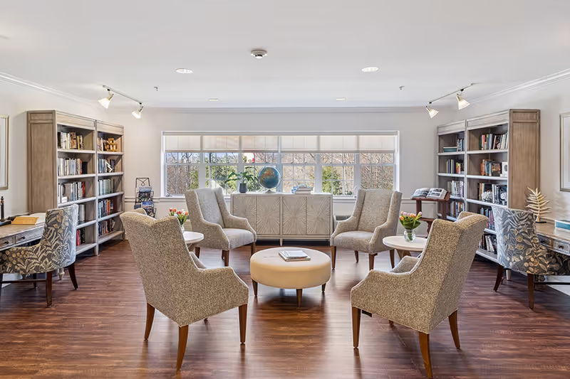 A bright and spacious senior living common area with six upholstered armchairs arranged in a circle around a round ottoman. There are two small round tables with flower vases, bookshelves filled with books on either side, and two desks with patterned chairs. A large window at the back lets in natural light, and the room has wooden flooring and white walls.