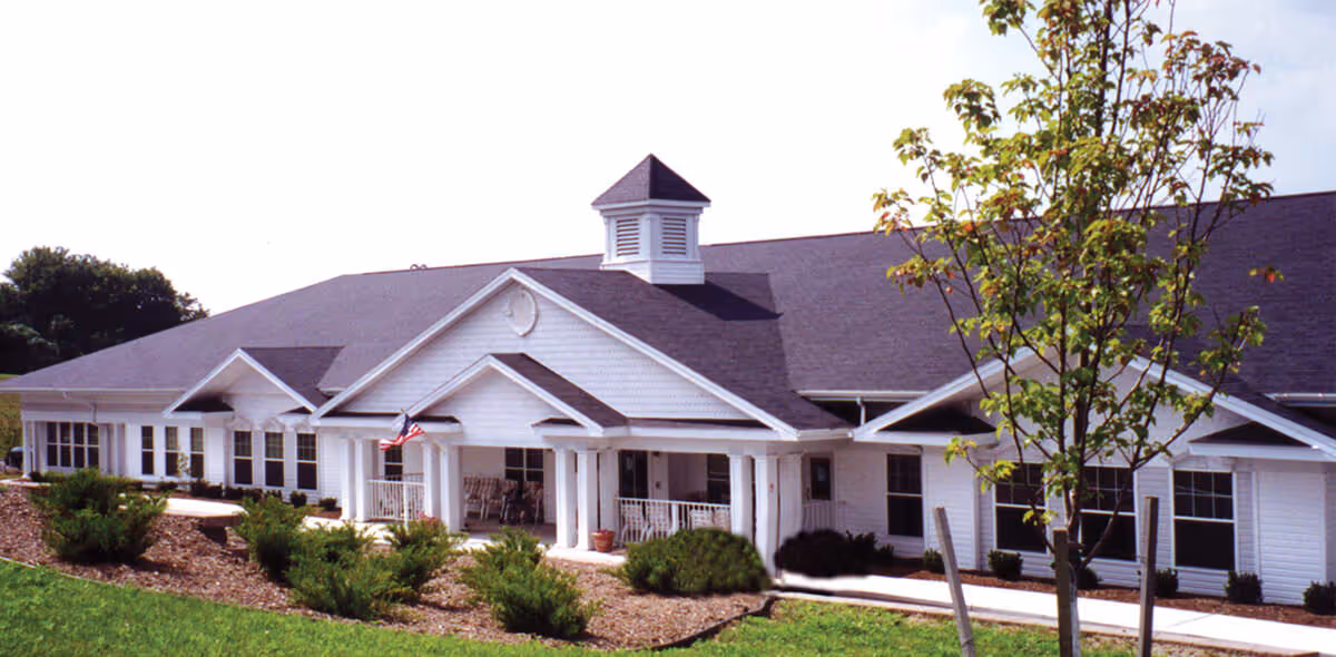 Exterior view of a single-story white building with a dark roof, featuring a covered porch with rocking chairs, an American flag, and landscaped bushes and trees in front.