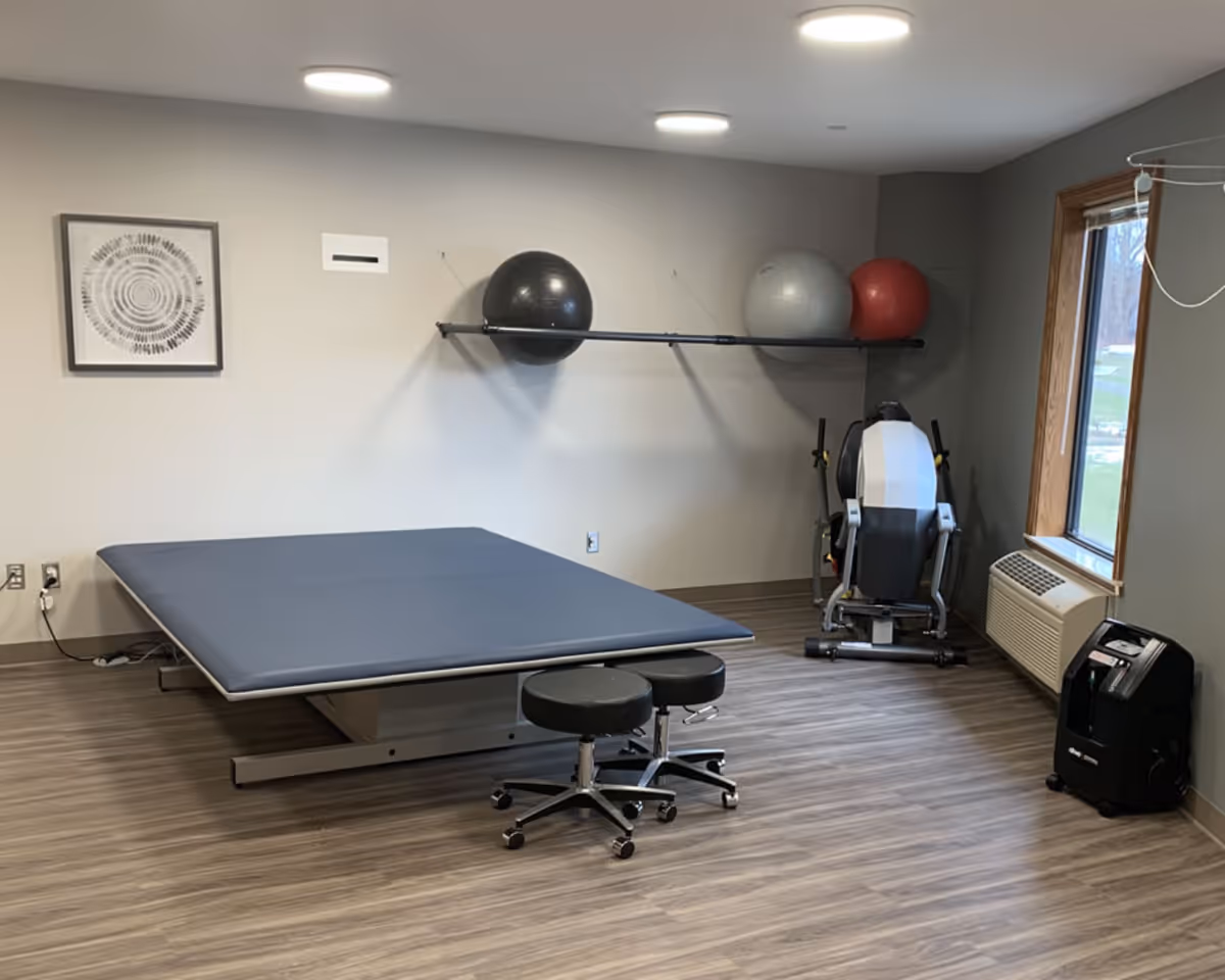 A therapy/exercise room with a padded treatment table, two rolling stools, exercise balls on a wall rack, and a piece of exercise equipment by a window.