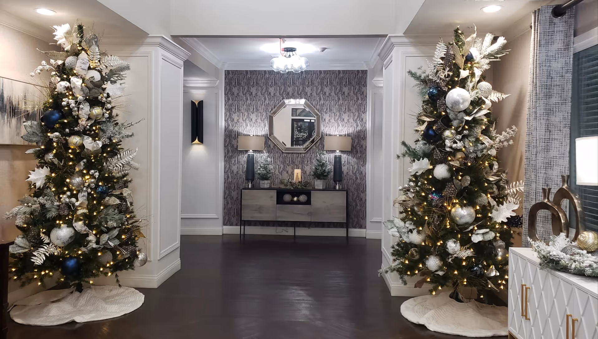 Interior hallway decorated for the holidays with two Christmas trees adorned with white, silver, and blue ornaments and lights. The hallway features white paneled walls, a dark wood floor, a console table with two lamps and decorative plants, and a geometric mirror on a textured accent wall.