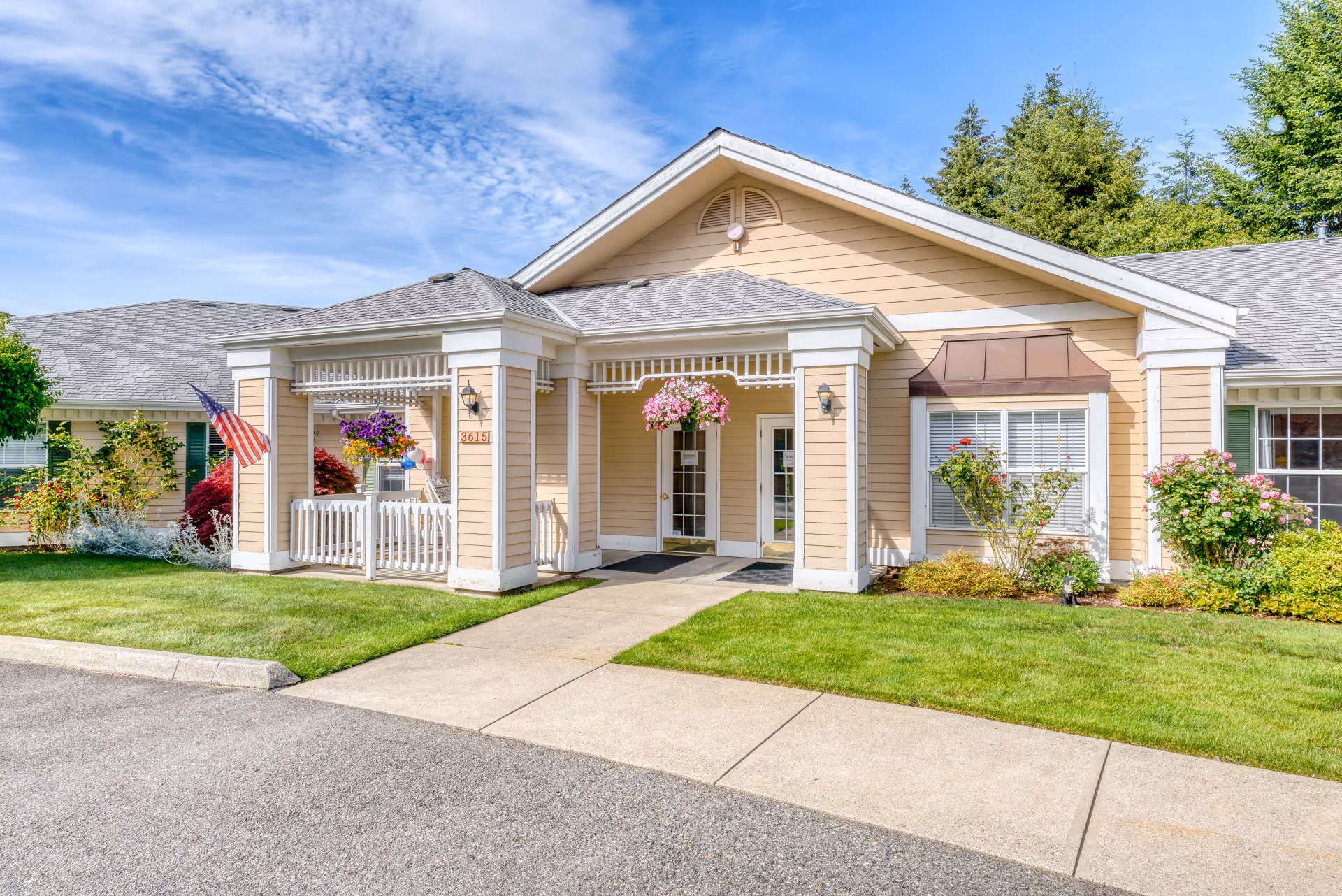 Front entrance of a single-story beige memory care building with a covered porch, hanging flower baskets, and an American flag.