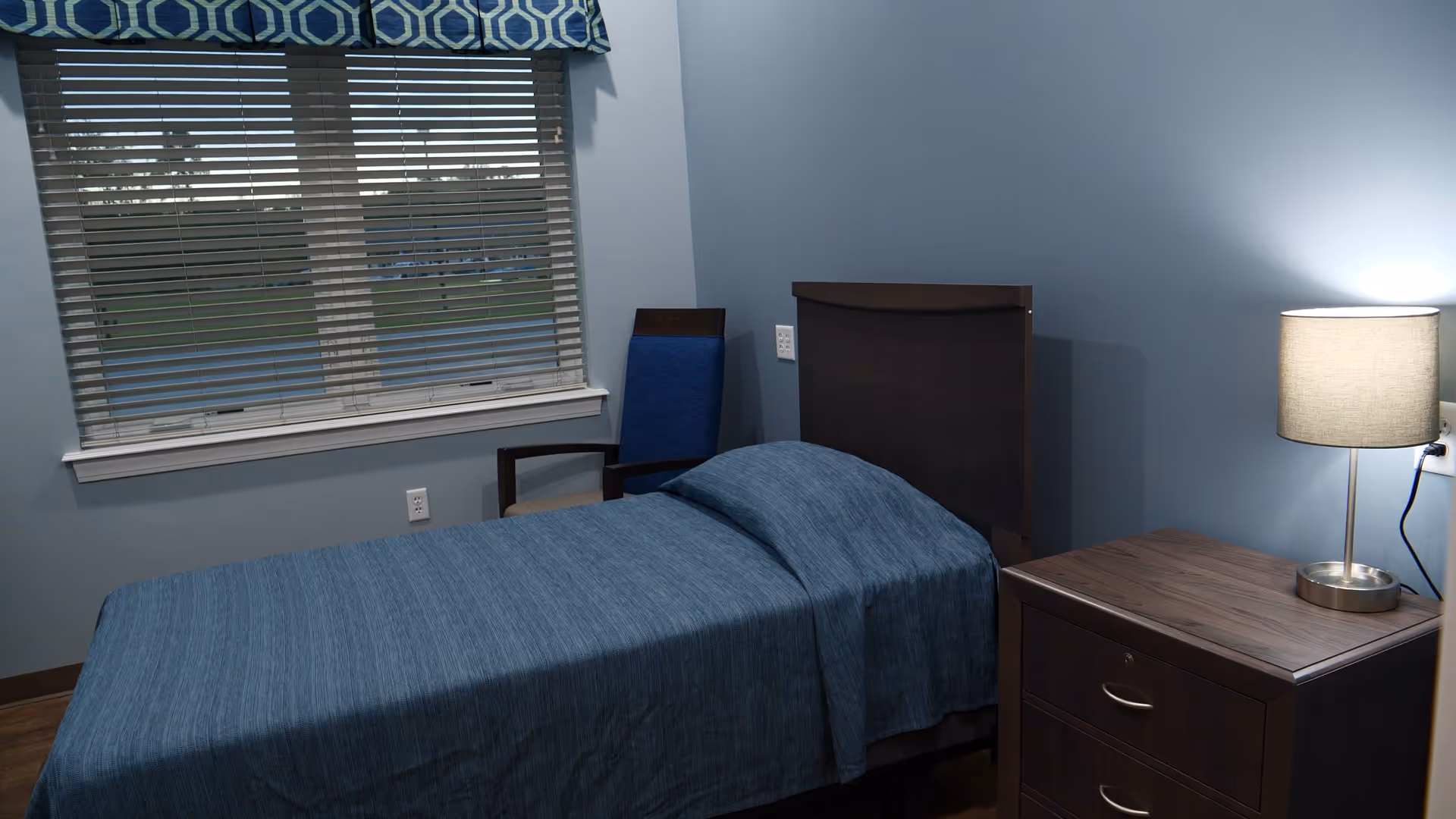 A single bed with blue bedding in a furnished bedroom featuring a wooden nightstand with a lamp, a chair, and a window with blinds.
