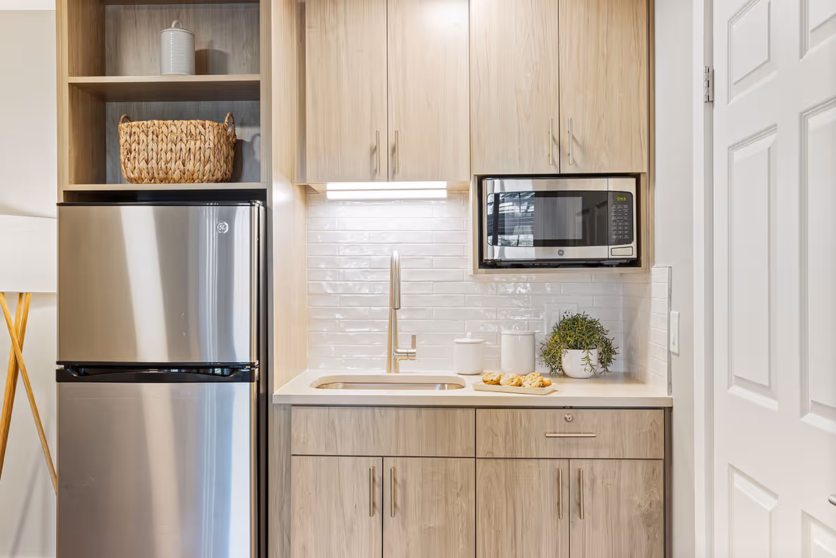 A modern kitchenette with light wood cabinets, a stainless steel refrigerator, a built-in microwave, a sink with a faucet, white subway tile backsplash, and a countertop with two white canisters, a small potted plant, and a plate of cookies.