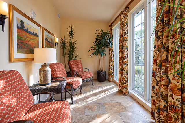 A sunlit sitting area with three red patterned armchairs arranged along a beige wall. A small table with a lamp is placed between two chairs. Two framed landscape paintings hang on the wall above the chairs. Large windows with floral curtains allow natural light to fill the space, and two tall potted plants are positioned near the corner.