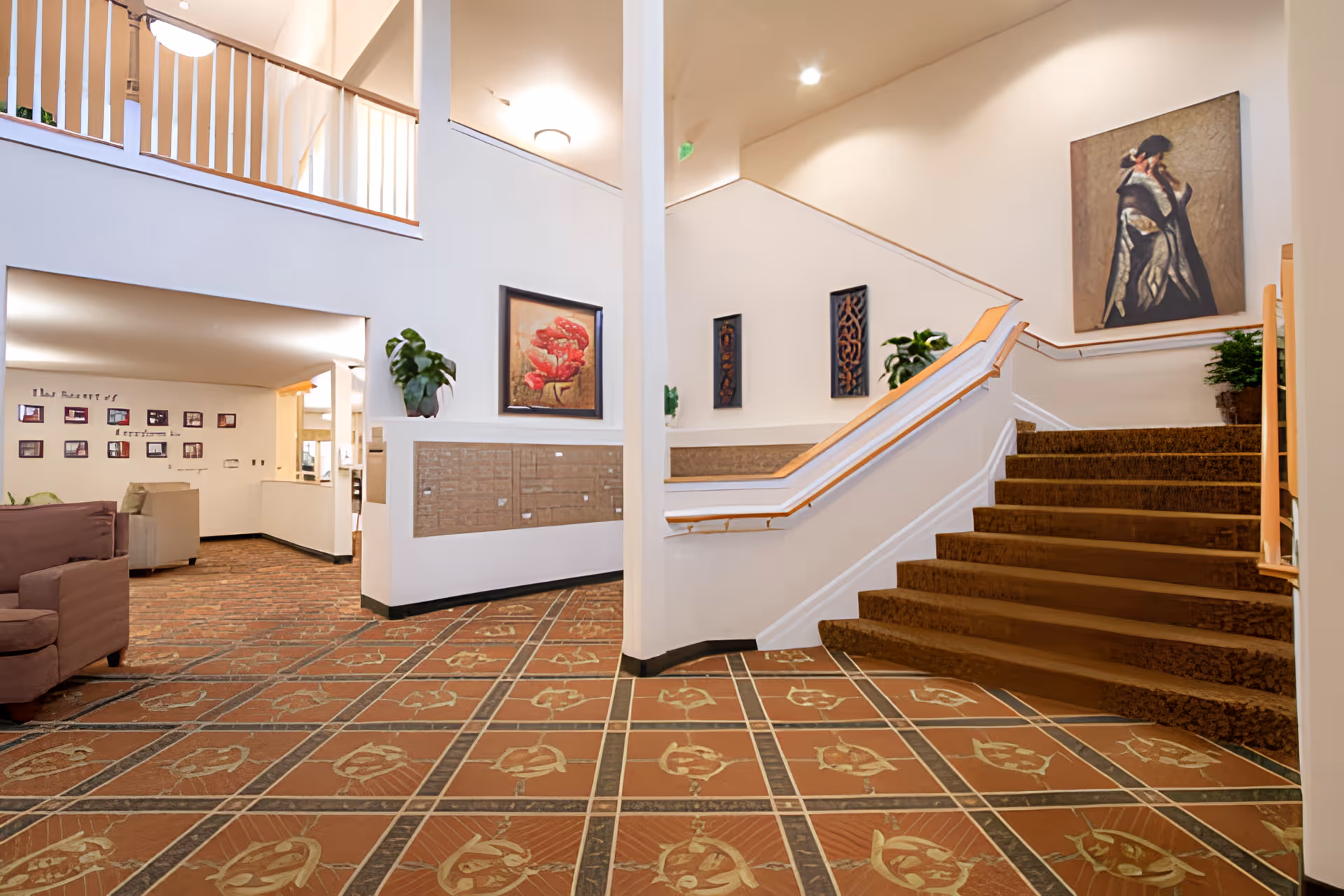 Interior view of a senior living facility lobby with patterned tile flooring, a staircase with carpeted steps and wooden handrails, several framed artworks on the walls, potted plants, and seating areas with armchairs.