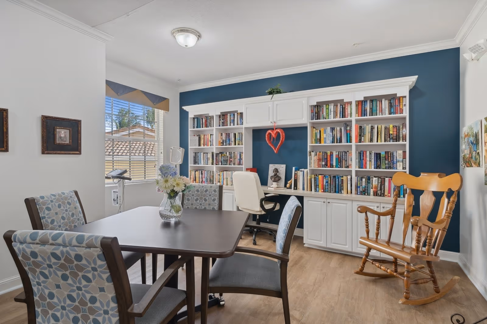 A cozy reading and activity room with a dark wooden table surrounded by four patterned chairs. A vase with flowers is placed on the table. In the background, there is a white built-in bookshelf filled with books against a dark blue wall. A white office chair is positioned at the desk area of the bookshelf. A wooden rocking chair is placed to the right. The room has light wood flooring and a window with blinds letting in natural light.