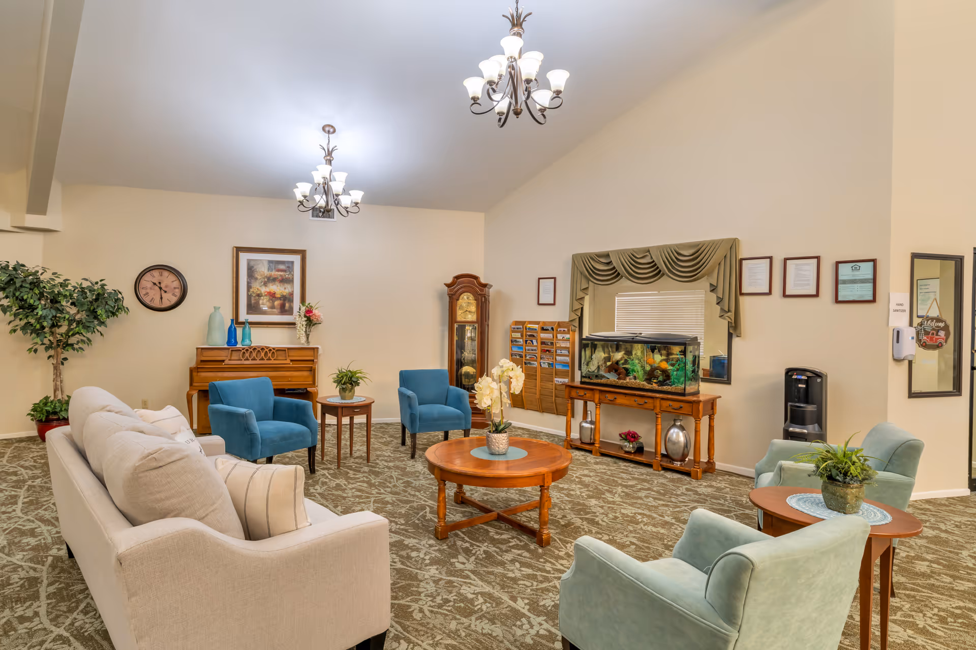 A cozy living room area in a senior living facility featuring a beige sofa, several blue and green armchairs, a round wooden coffee table with a potted plant, a piano with decorative vases and flowers, a grandfather clock, a large fish tank on a wooden stand, framed pictures and certificates on the walls, and two chandeliers hanging from the ceiling.