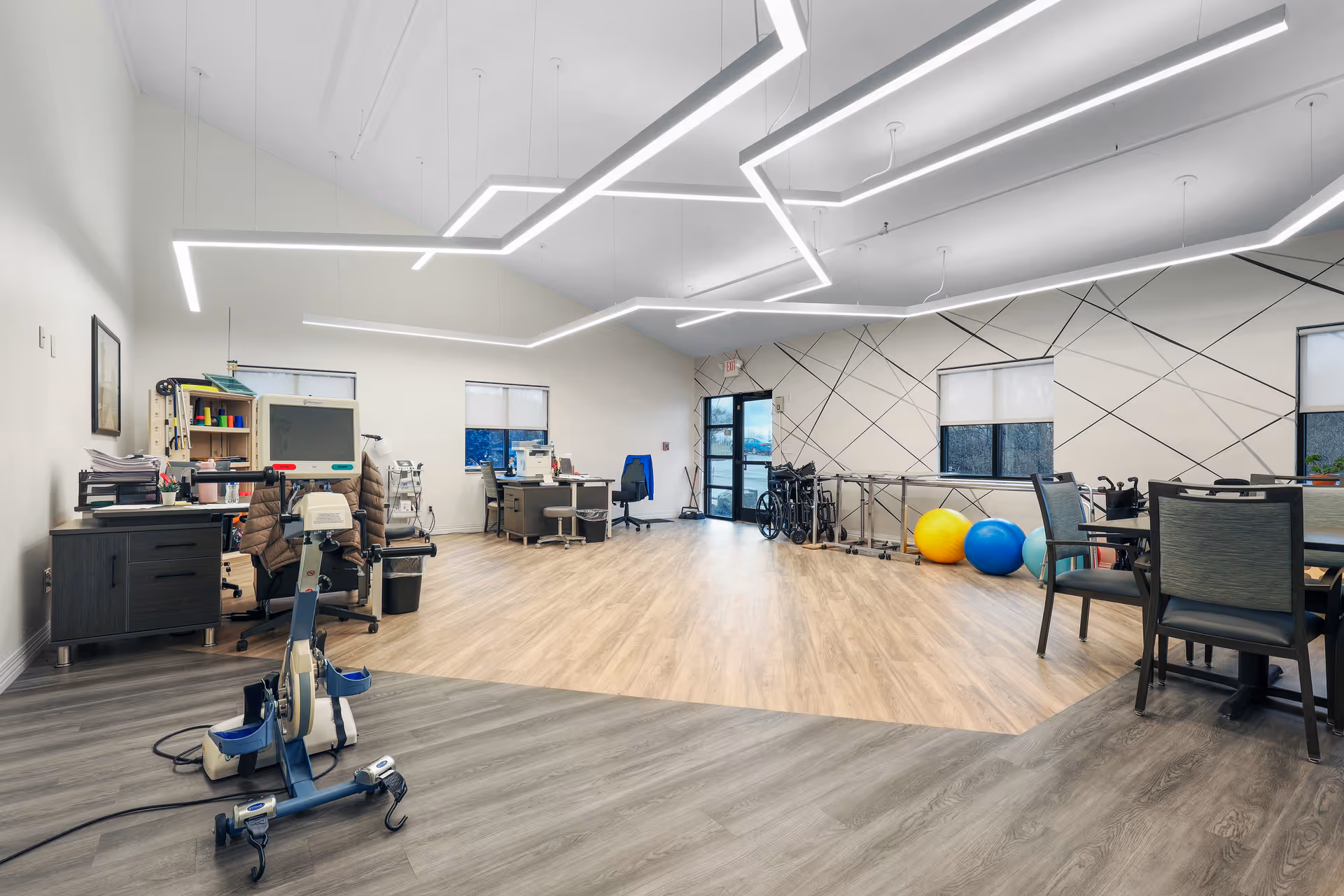A spacious, well-lit rehabilitation or therapy room with modern geometric ceiling lights, exercise equipment including a stationary bike, therapy balls, wheelchairs, desks with office chairs, and a patterned accent wall with windows.