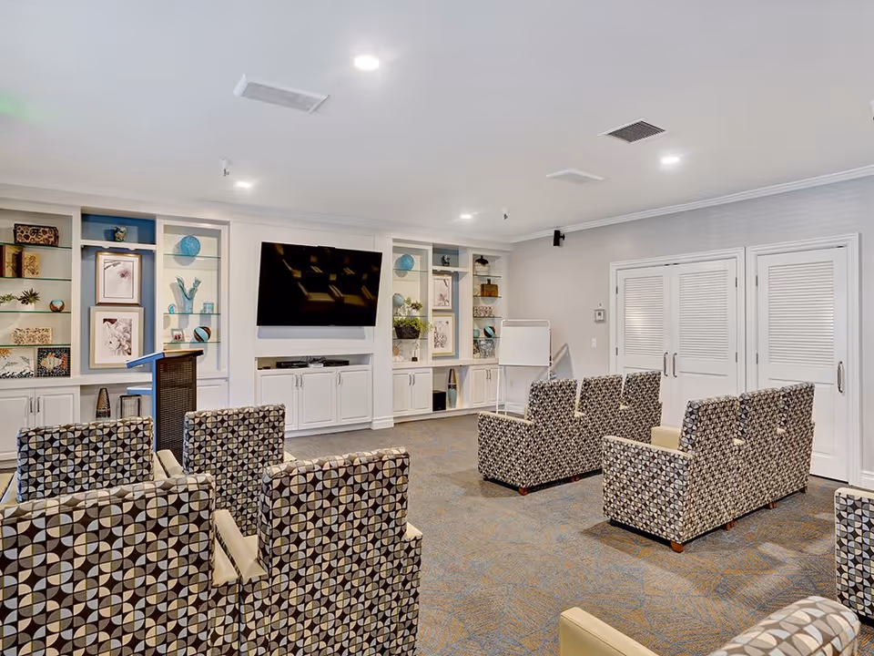 A well-lit common room with patterned armchairs arranged in rows facing a large flat-screen TV mounted on a white built-in cabinet wall with shelves displaying decorative items and framed artwork. There is a podium and a whiteboard on a stand near the back wall with double doors.