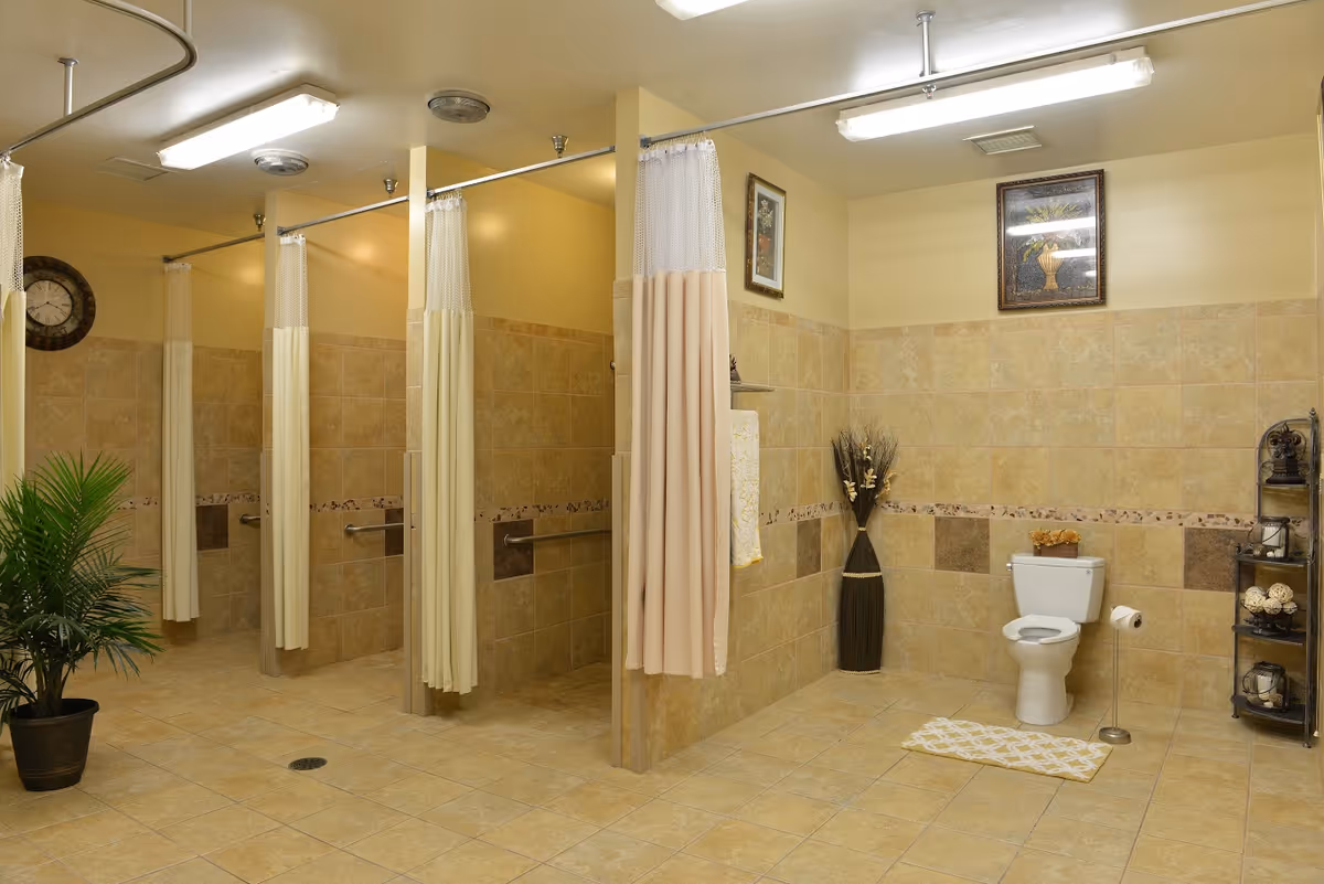 A spacious, clean bathroom area with multiple shower stalls separated by beige curtains. The walls and floor are tiled in light brown tones. There is a white toilet with a toilet paper holder and a small rug in front of it. Decorative elements include a potted plant, a tall vase with dried flowers, framed artwork on the walls, and a small shelving unit with decorative items.