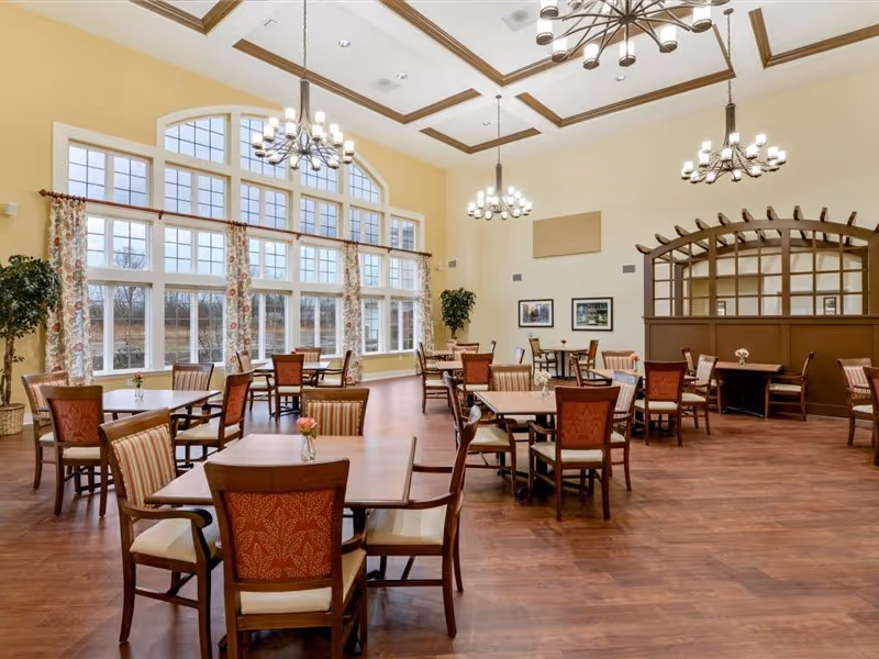 A spacious dining room with multiple wooden tables and chairs arranged neatly. Large windows with floral curtains allow natural light to fill the room. The ceiling features decorative beams and multiple chandeliers. There are framed pictures on the walls and a wooden lattice partition in the background.