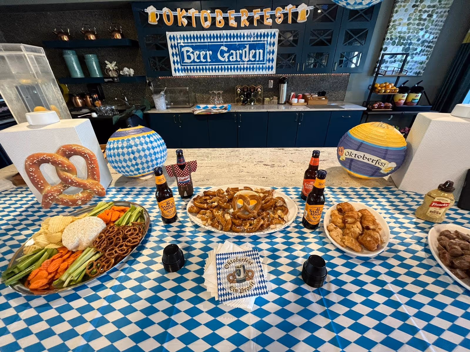 Oktoberfest-themed buffet table with pretzels, snacks, beer bottles and decorations in a communal dining area.