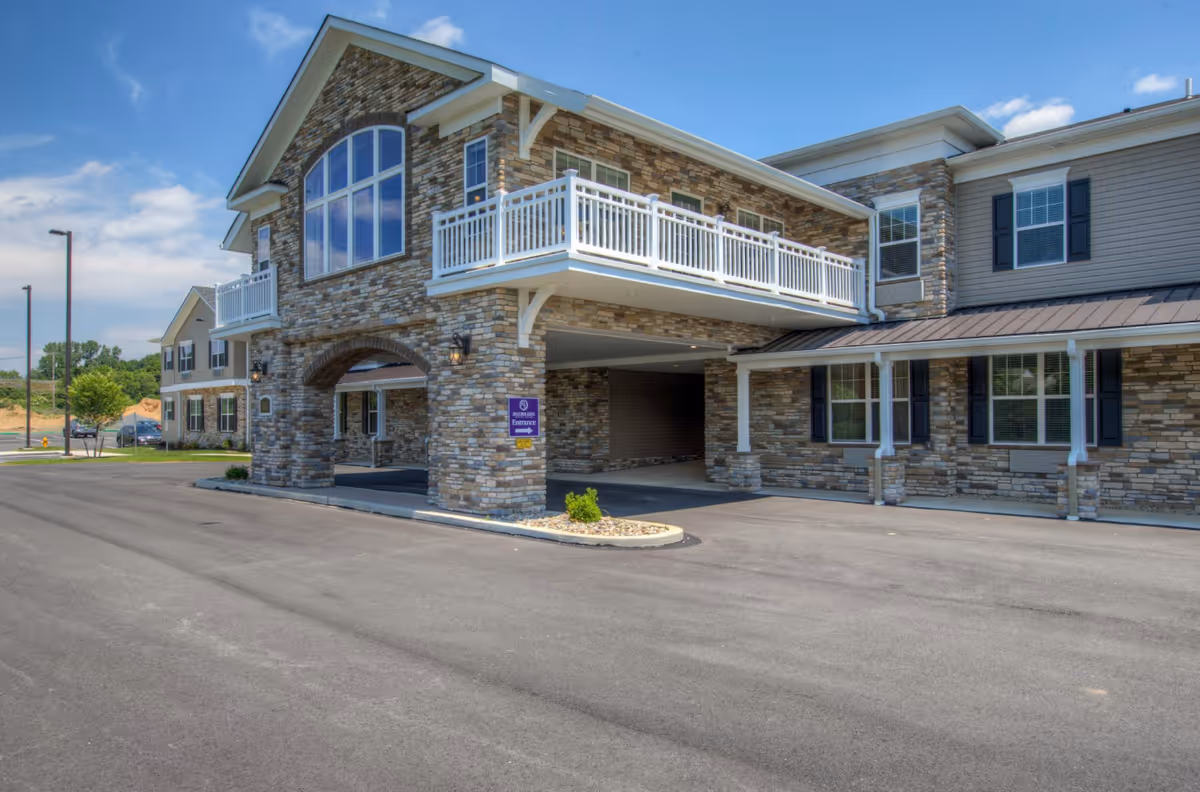Stone-clad senior living facility entrance with a covered porte-cochère, balcony, and paved driveway under a blue sky.