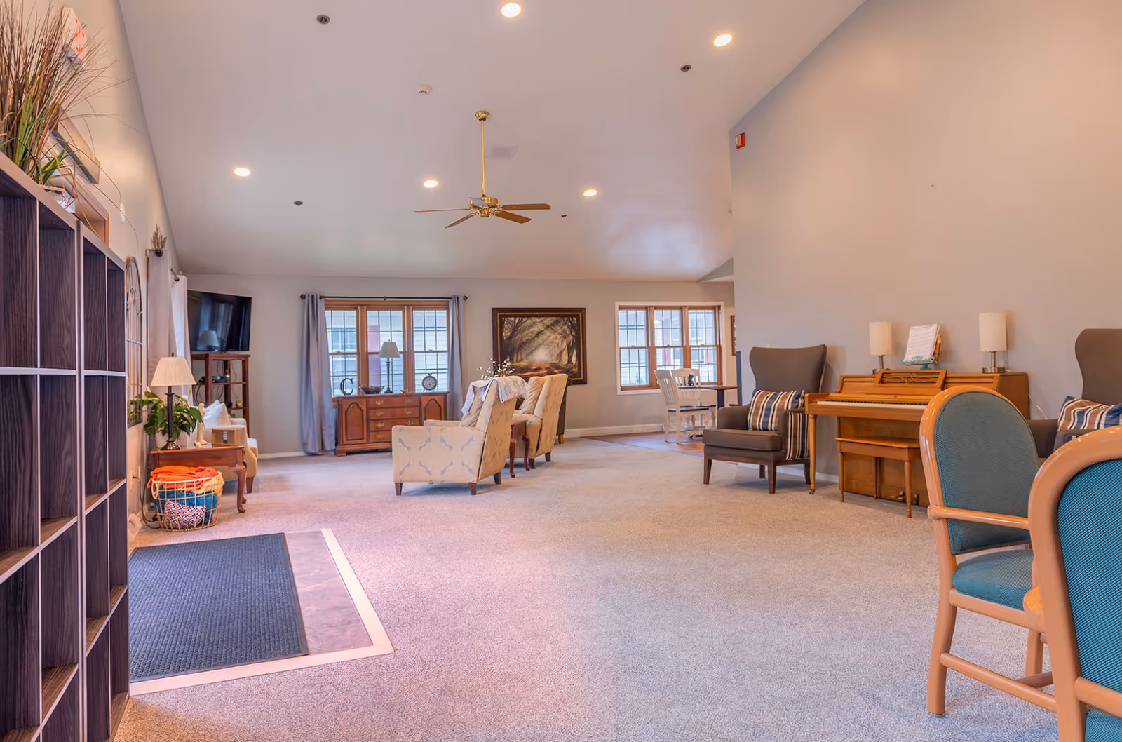 Open, well-lit senior living common room with seating, a piano, TV, and large windows.