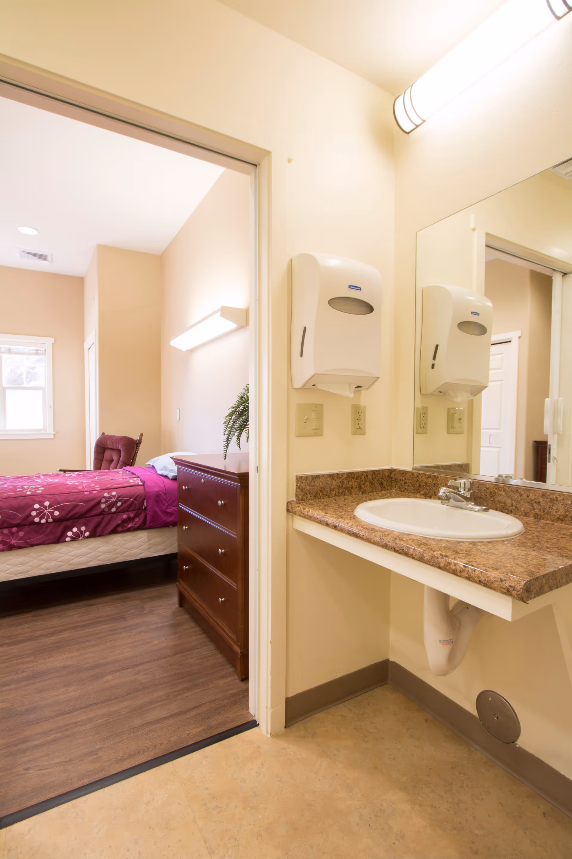 View of a bedroom with a bed covered in a purple floral bedspread and a wooden dresser, seen through an open doorway from a small area with a sink, countertop, and wall-mounted paper towel dispenser.