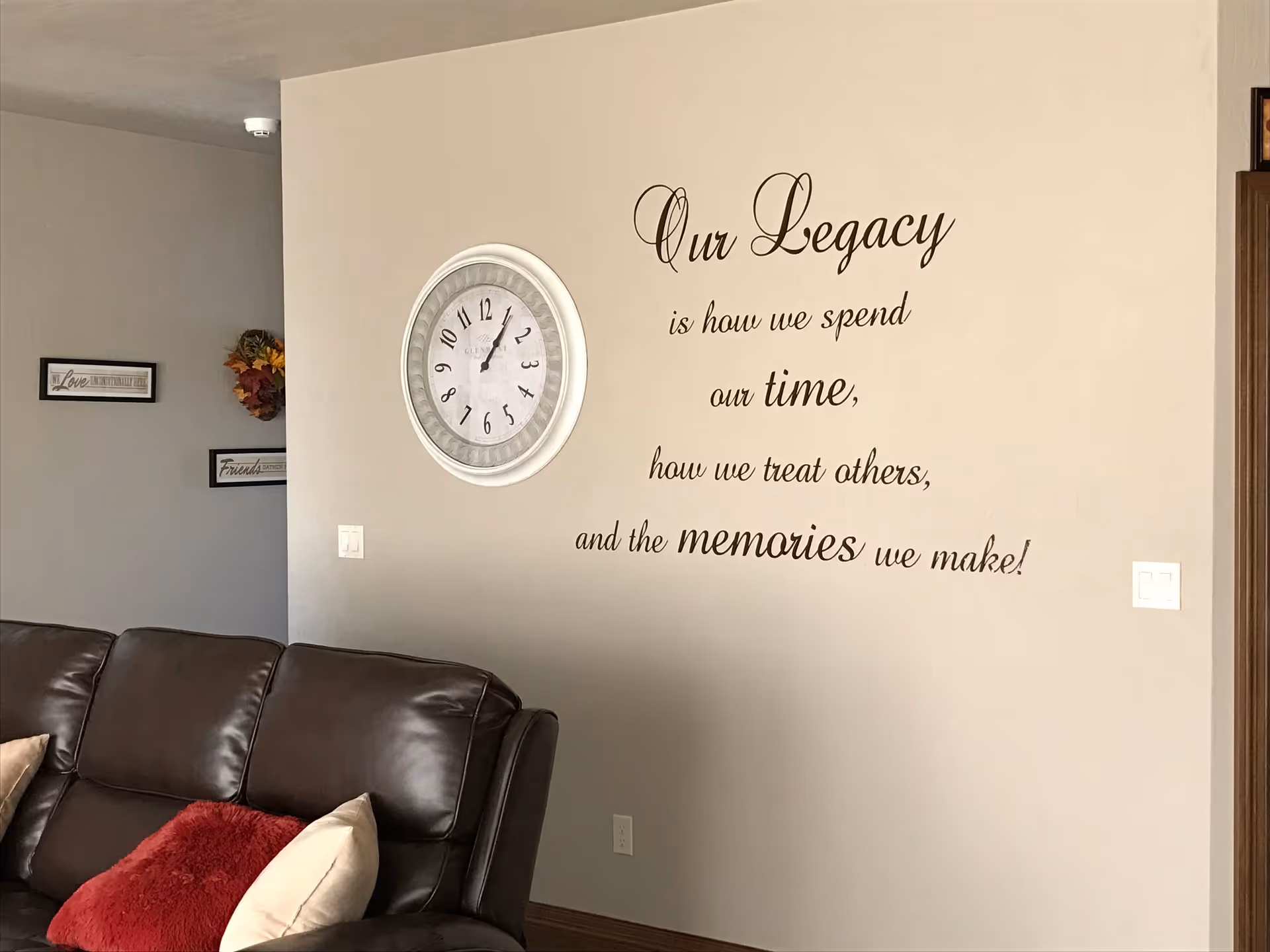 Interior view of a living room area with a brown leather couch adorned with beige and red pillows. On the beige wall above the couch is a large round clock and a decorative wall quote that reads: 'Our Legacy is how we spend our time, how we treat others, and the memories we make!'. There are also two framed signs and a floral wreath on the adjacent wall.